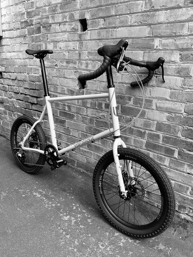 White small-wheeled bike with drop bars against a brick wall