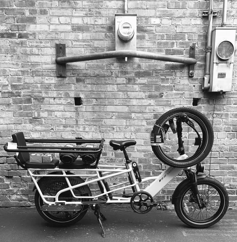White electric cargo bike with a spare wheel carried on the front, against a brick wall